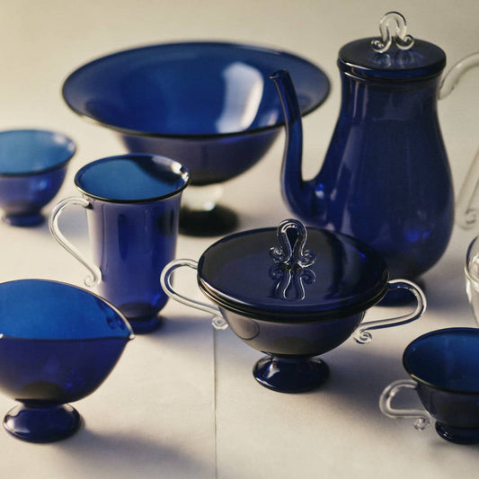 Collection of blue glassware including cups, bowls, and a teapot on a neutral background