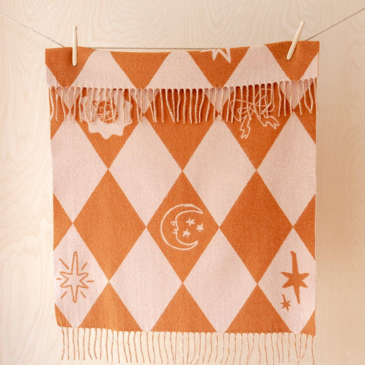 An orange and cream harlequin patterned throw blanket with fringe, hanging from clips on a clothesline against a light wood paneled wall.