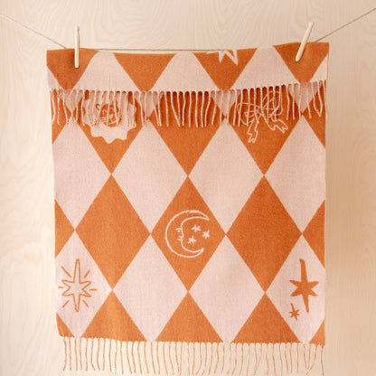 An orange and cream harlequin patterned throw blanket with fringe, hanging from clips on a clothesline against a light wood paneled wall.