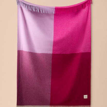 A square woven throw blanket with fringe on three sides, featuring a four-quadrant color-block pattern in shades of lavender, magenta, and deep burgundy, displayed against a solid beige wall.