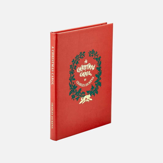 A hardcover book of 'A Christmas Carol' by Charles Dickens, featuring a vibrant red cover with a green holly wreath and gold foil lettering, shown on a clean white background.
