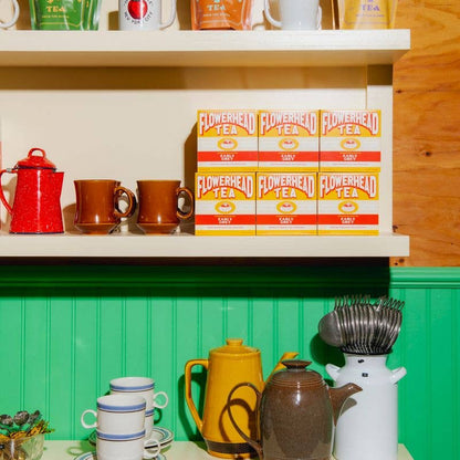 A styled tea display featuring multiple packages of Flowered Tea, including boxes and bags, arranged on white shelves alongside ceramic mugs, teapots, and utensils against a green and cream paneled background.