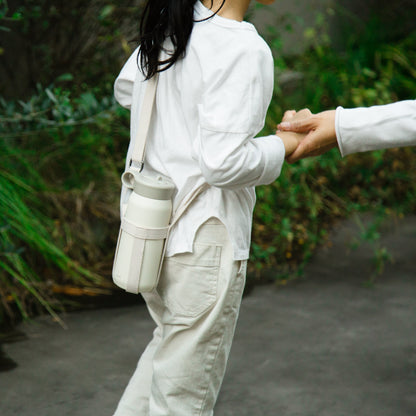 Person wearing a white outfit with a beige bag, standing outdoors with greenery in the background.