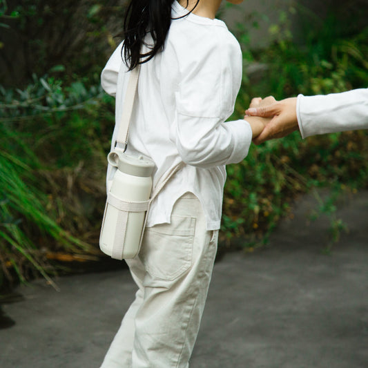 Person wearing a white outfit with a beige bag, standing outdoors with greenery in the background.