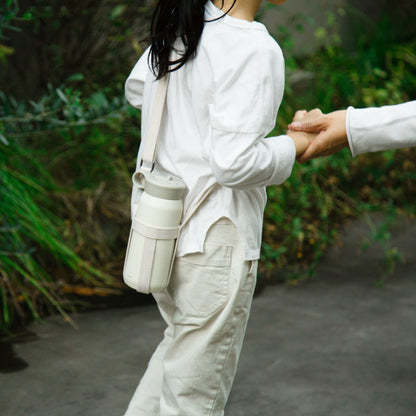 Person wearing a white shirt and beige pants with a beige bag, standing outdoors.
